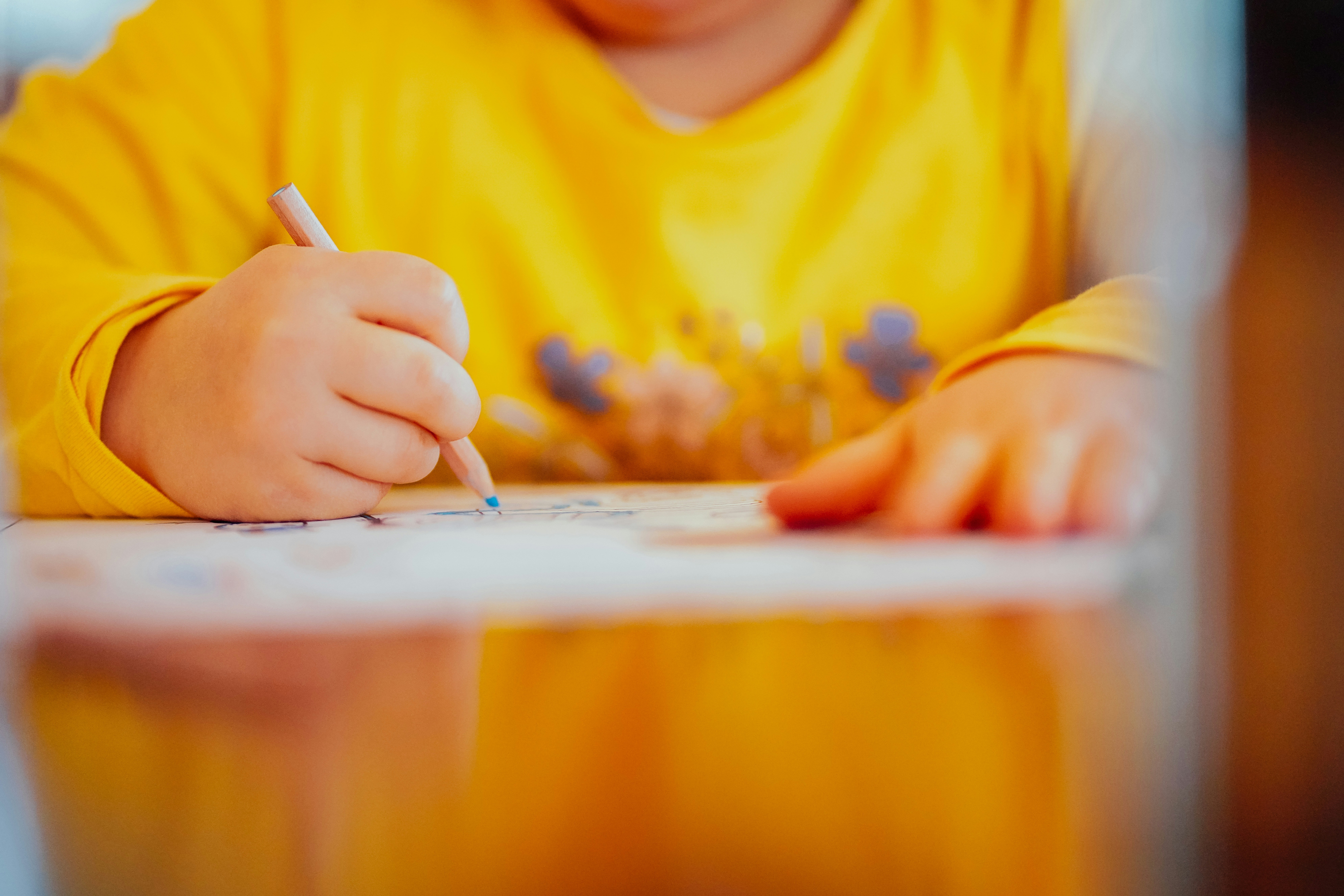 A child wearing a yellow long-sleeve shirt holds a pencil and draws on a piece of paper at a table, shown in a close-up view of the child's hands and upper body.