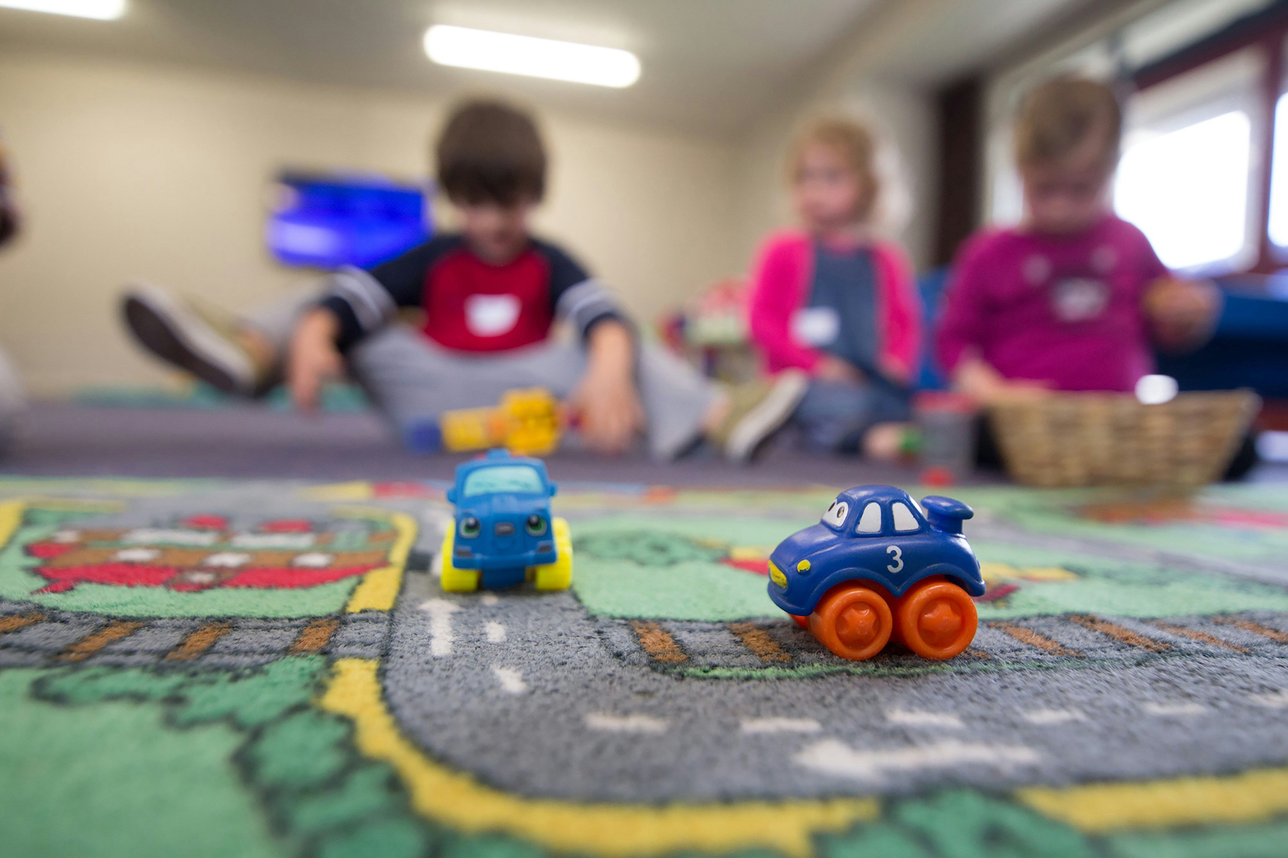 Toy cars placed on a colorful road-patterned play mat in a childcare room, with several young children sitting on the floor in the background.