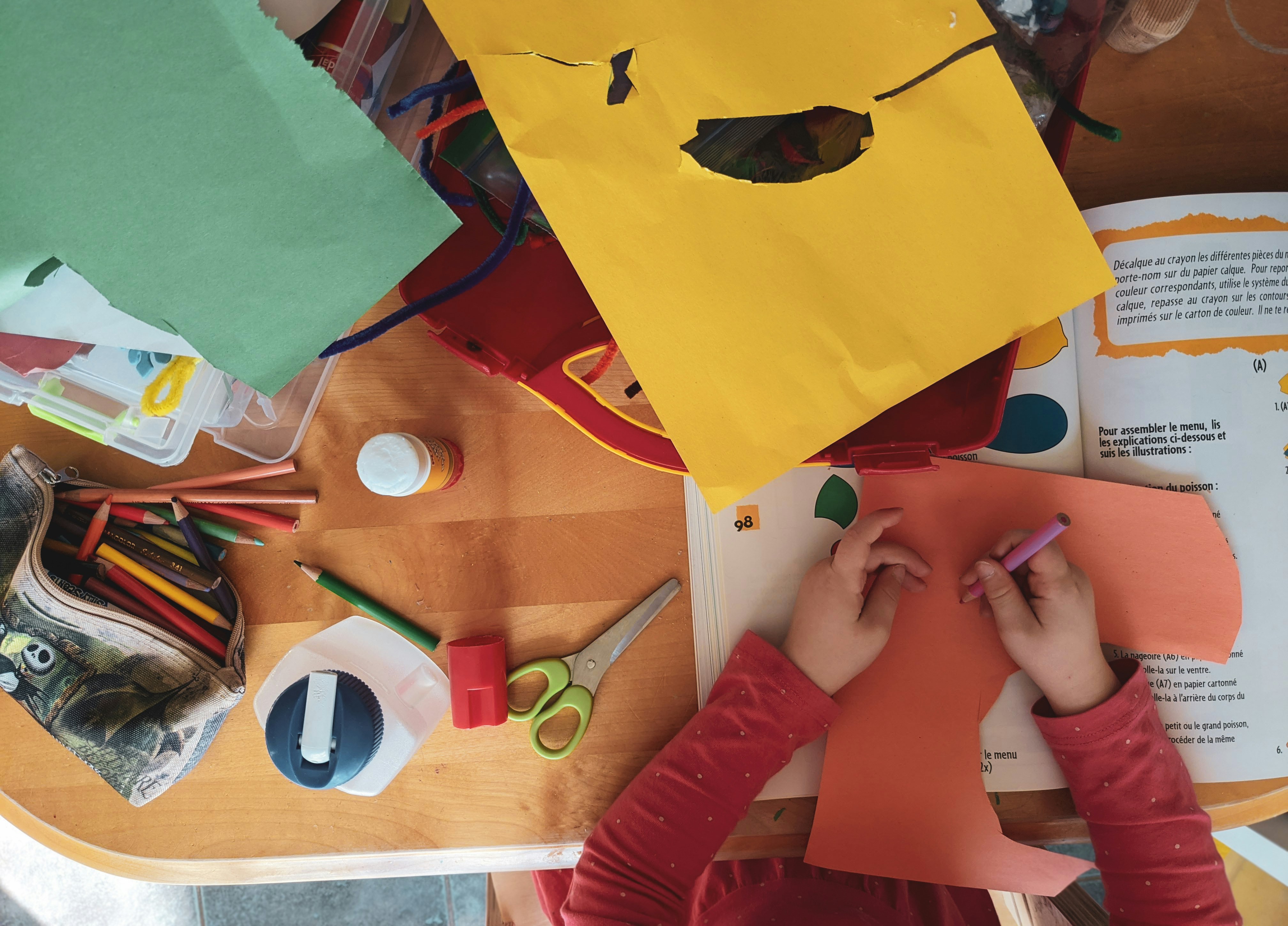 A child’s hands work on an orange piece of paper during an arts and crafts activity, surrounded by colored paper, markers, scissors, glue, and other craft supplies spread across the table.