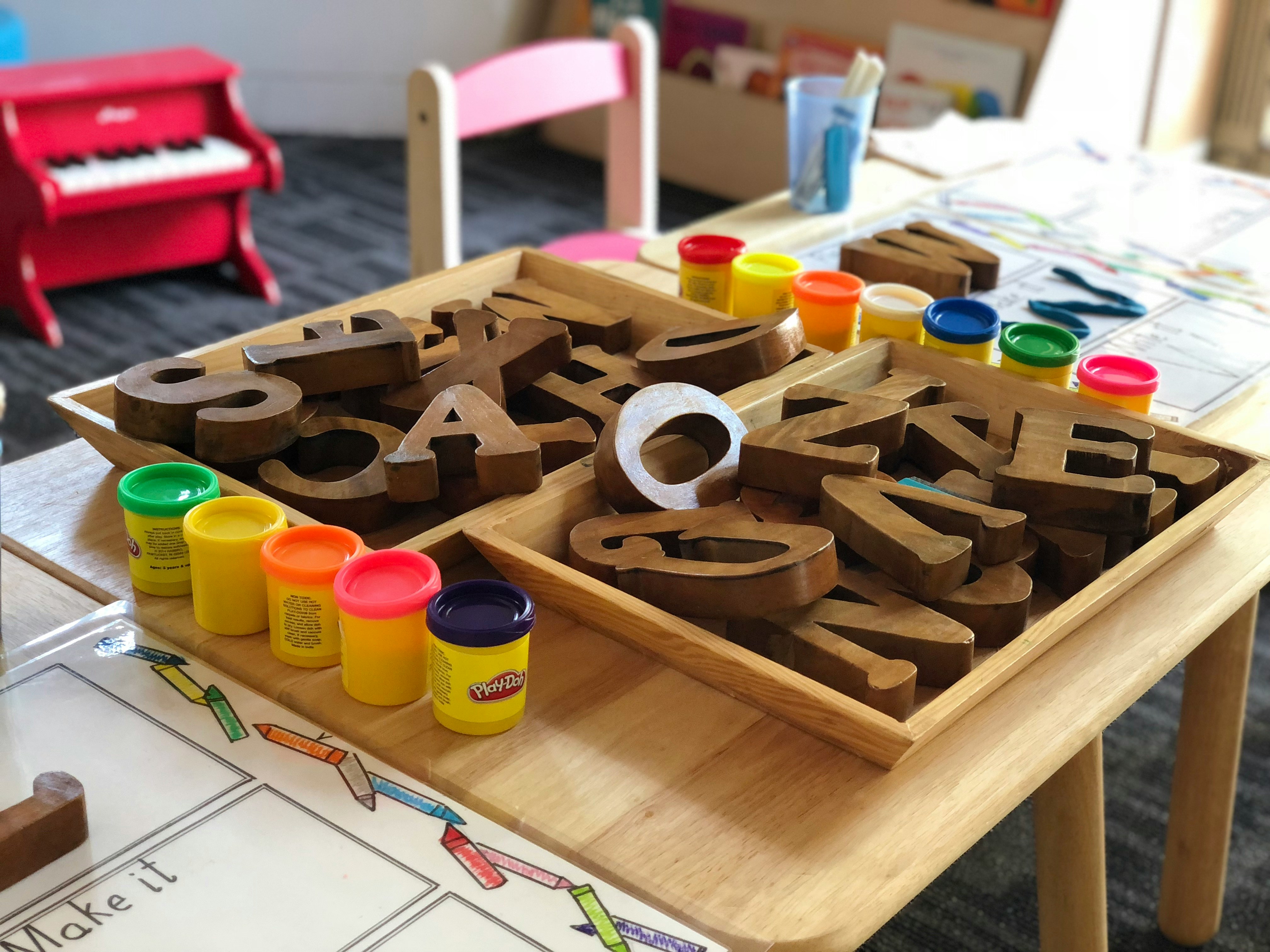 A table set up with wooden alphabet letters in sorting trays, alongside several containers of colorful Play-Doh and children's activity sheets.