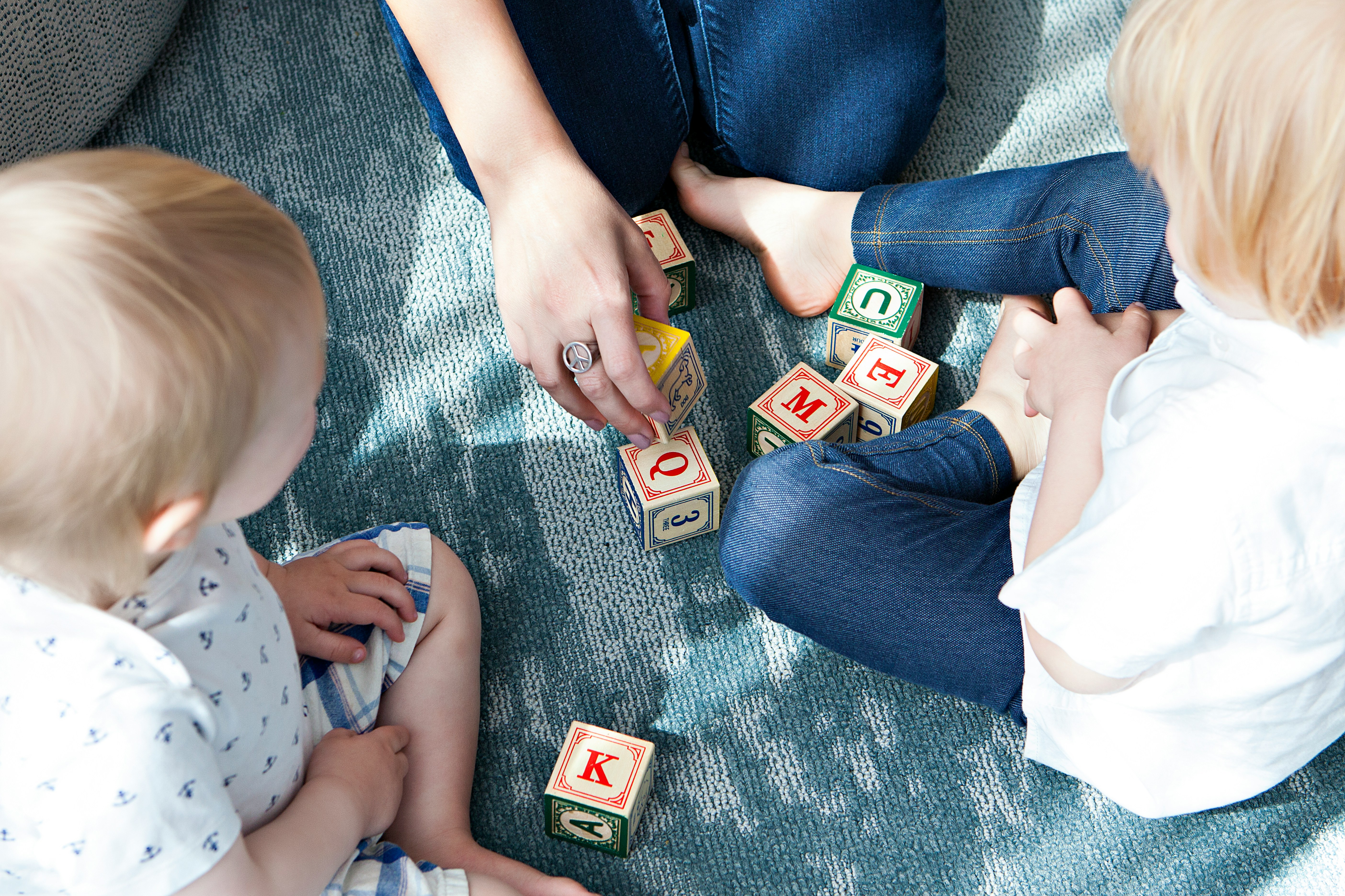 Two young children sit on a carpeted floor playing with wooden alphabet blocks, with an adult seated nearby helping arrange the blocks.