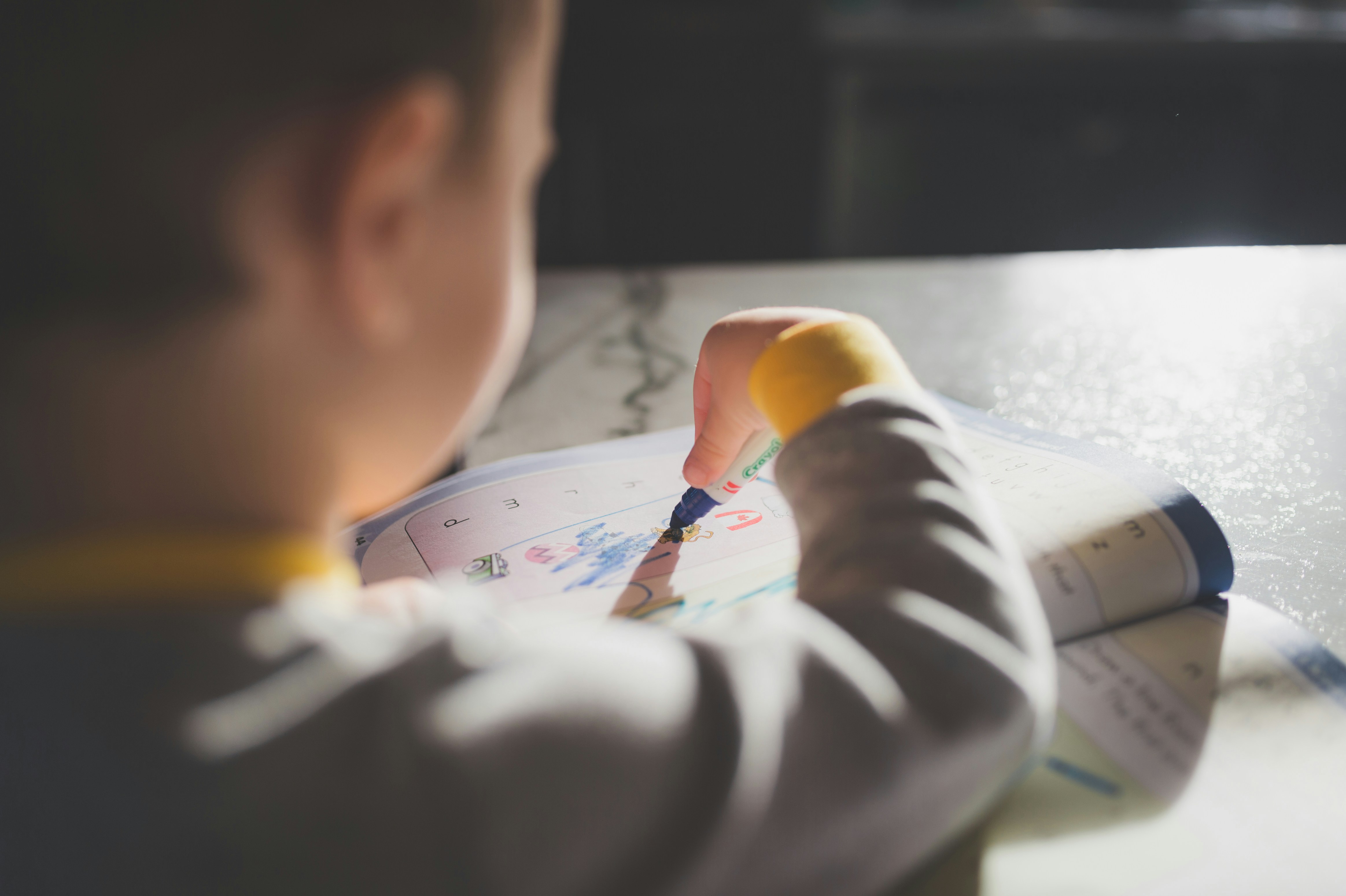 A young child sits at a table coloring in a picture with a blue marker, with sunlight shining across the page from the left.